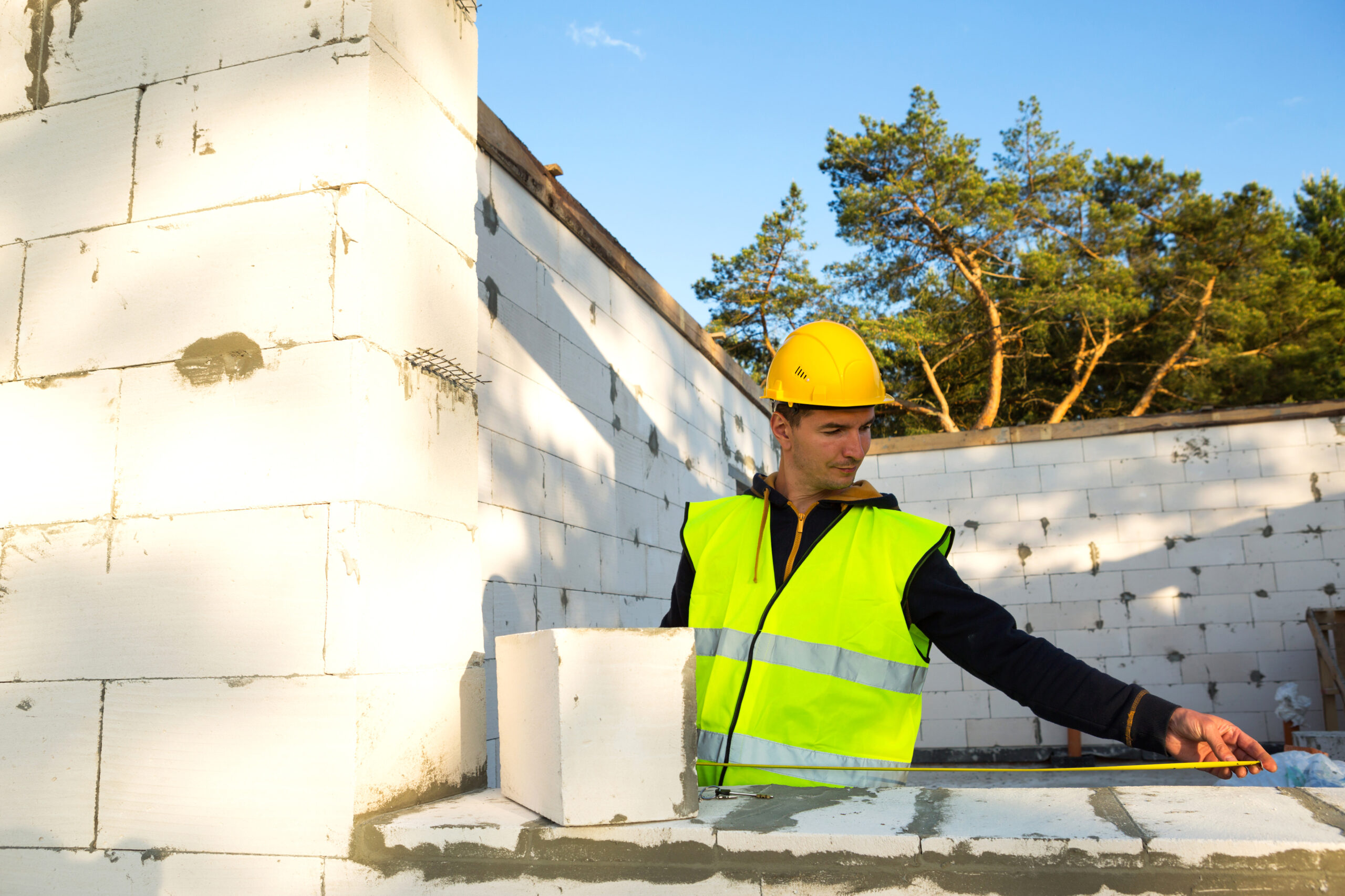 Construction worker at construction site measures the length of the window opening and the wall with tape measure. Cottage are made of porous concrete blocks, protective clothing - hardhat and a vest