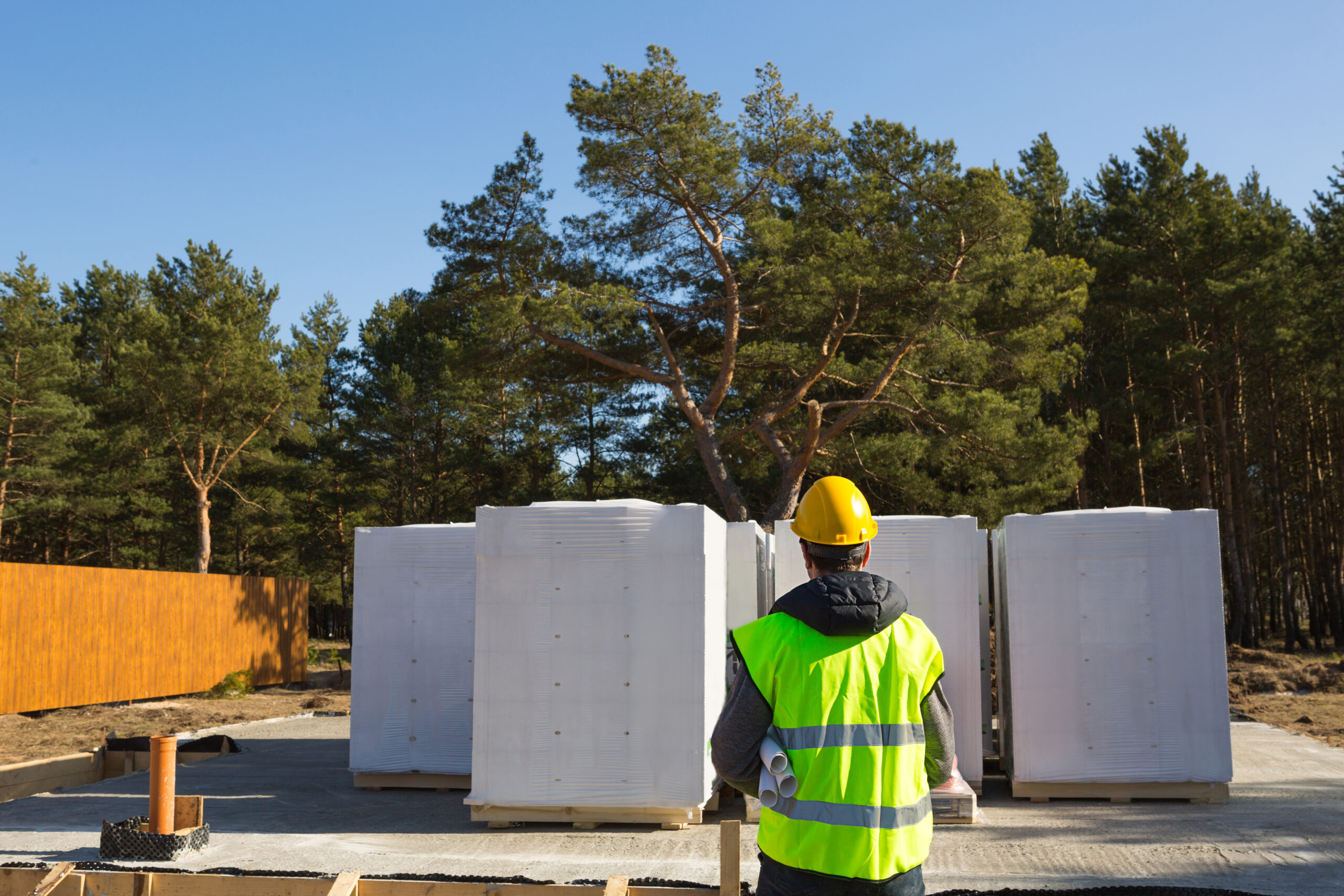 The project architect's back is on the construction site of a house with the foundation laid and the blocks delivered. Construction worker in a protective yellow helmet and a signal vest. Mock up
